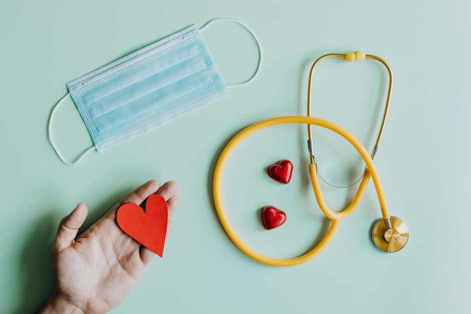Top view of crop anonymous person hand with red paper heart on table with stethoscope and medical mask for coronavirus prevention